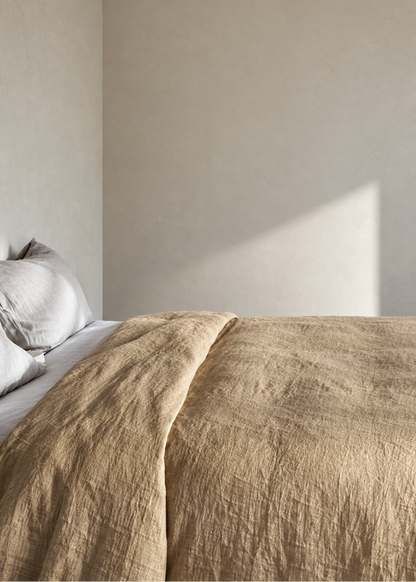 A close-up of a textured beige linen bedspread and matching pillowcases on a bed, set against a minimalist beige wall with soft natural light casting a shadow.