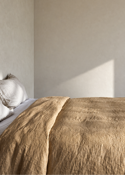 A close-up of a textured tan linen duvet cover spread across a bed with white pillows, set against a minimalist beige bedroom wall with soft sunlight casting a shadow.