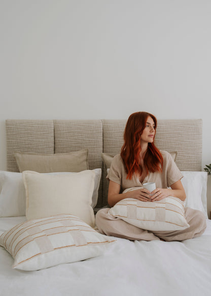 Woman sitting on a bed holding a mug, surrounded by pillows and a neutral headboard.