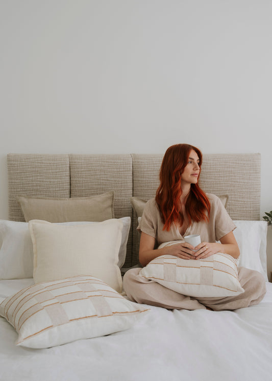 Woman sitting on a bed holding a mug, surrounded by pillows and a neutral headboard.