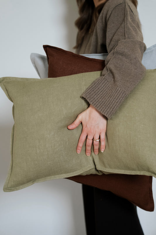 Person holding two pillows, one green and one brown, against a neutral background.
