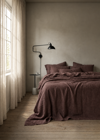 A rustic bedroom scene featuring a bed dressed in a textured, deep plum-brown linen bedding set, styled with matching pillows. The room has a warm, neutral plaster wall, light wood flooring, sheer curtains by a window, and a black wall lamp.