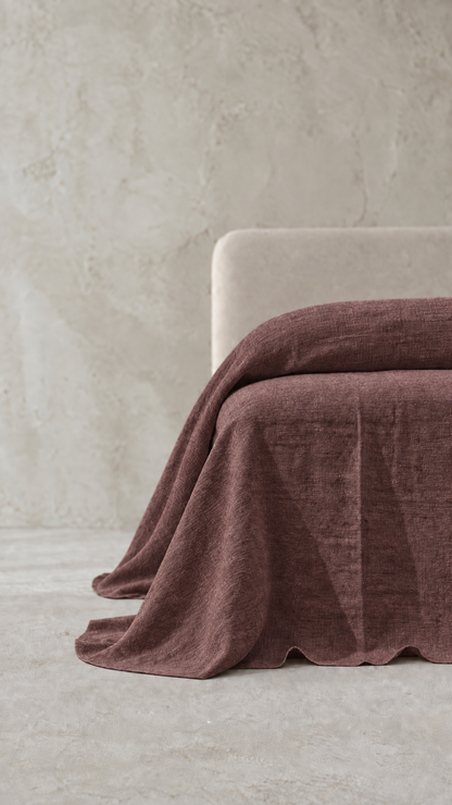 A draped dusty rose or mauve-colored textured throw blanket or bedspread, shown partially folded over the corner of a light beige upholstered chair in a minimalist room setting with a textured concrete wall and floor.