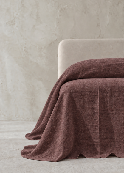 A draped dusty rose or mauve-colored textured throw blanket or bedspread, shown partially folded over the corner of a light beige upholstered chair in a minimalist room setting with a textured concrete wall and floor.