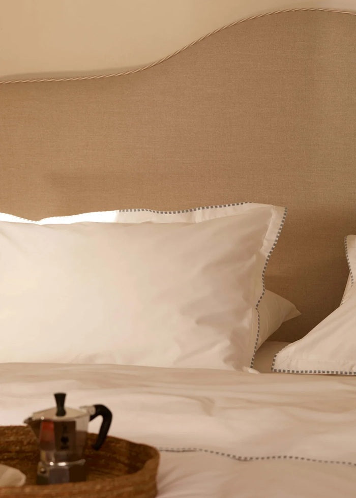 A close-up of a neatly made bed with white pillows and bedding, a beige upholstered headboard, and a small woven tray holding a metal coffee pot in the foreground.