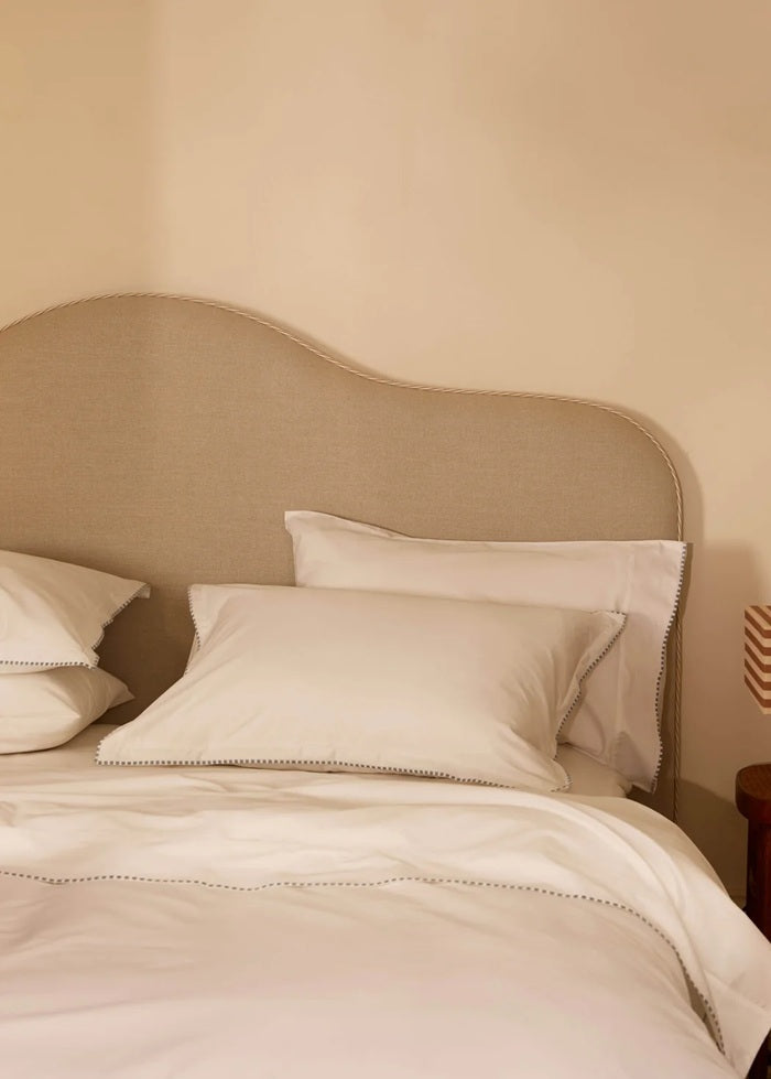 A neatly made bed with white pillows and duvet, set against a beige upholstered headboard and a warm beige wall. A small wooden side table with a striped lamp is visible on the right.