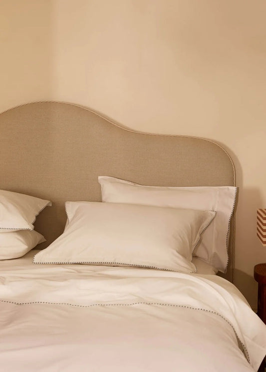 A neatly made bed with white pillows and duvet, set against a beige upholstered headboard and a warm beige wall. A small wooden side table with a striped lamp is visible on the right.