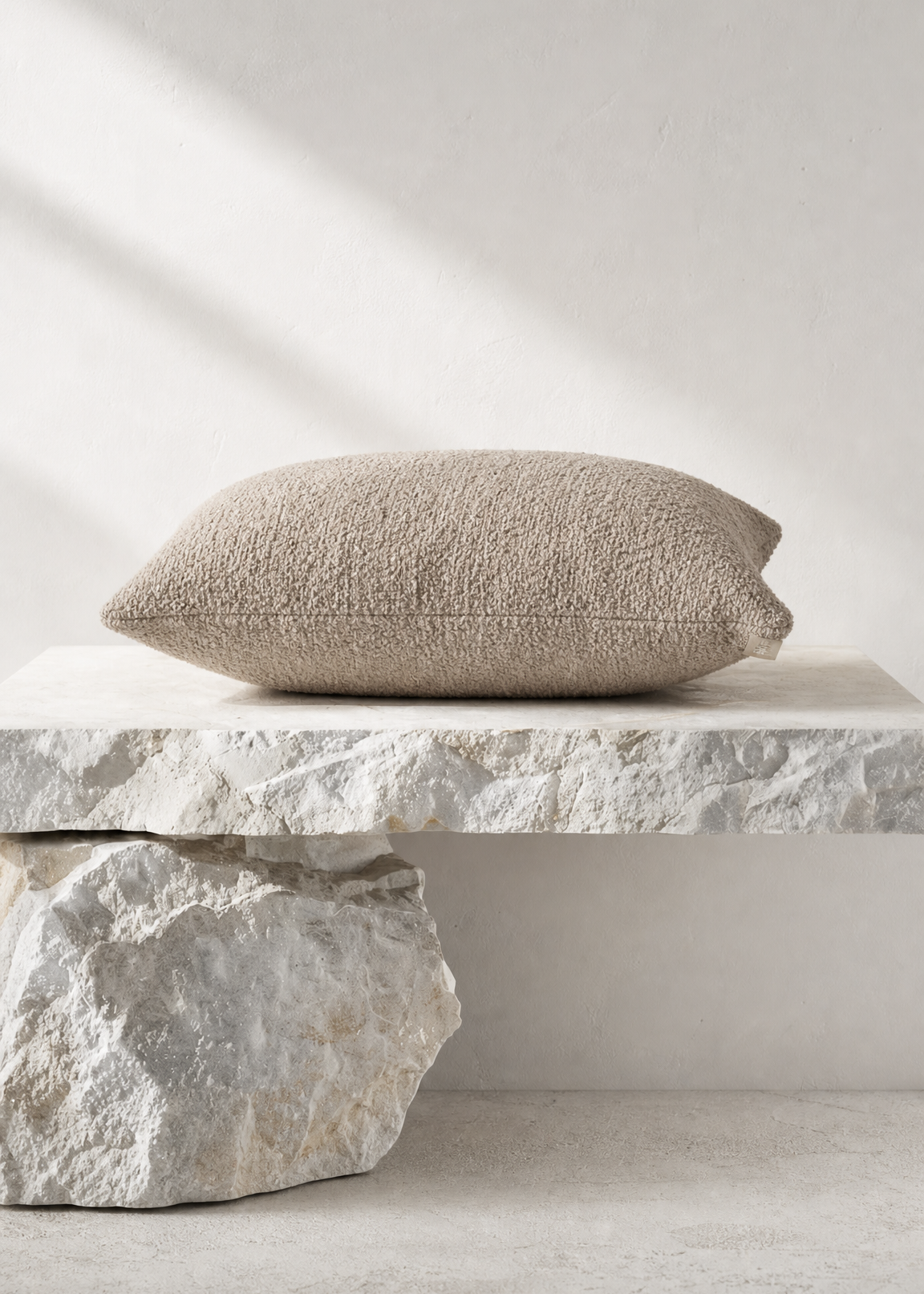 A textured beige throw pillow resting on a rustic stone pedestal against a neutral light gray wall with soft diagonal shadows.