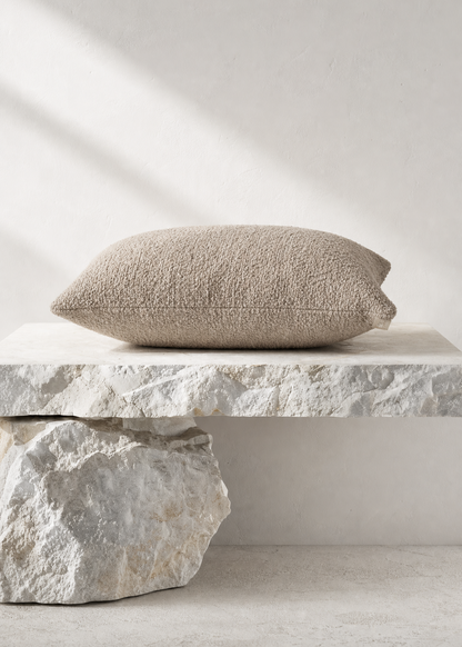 A textured beige throw pillow resting on a rustic stone pedestal against a neutral light gray wall with soft diagonal shadows.