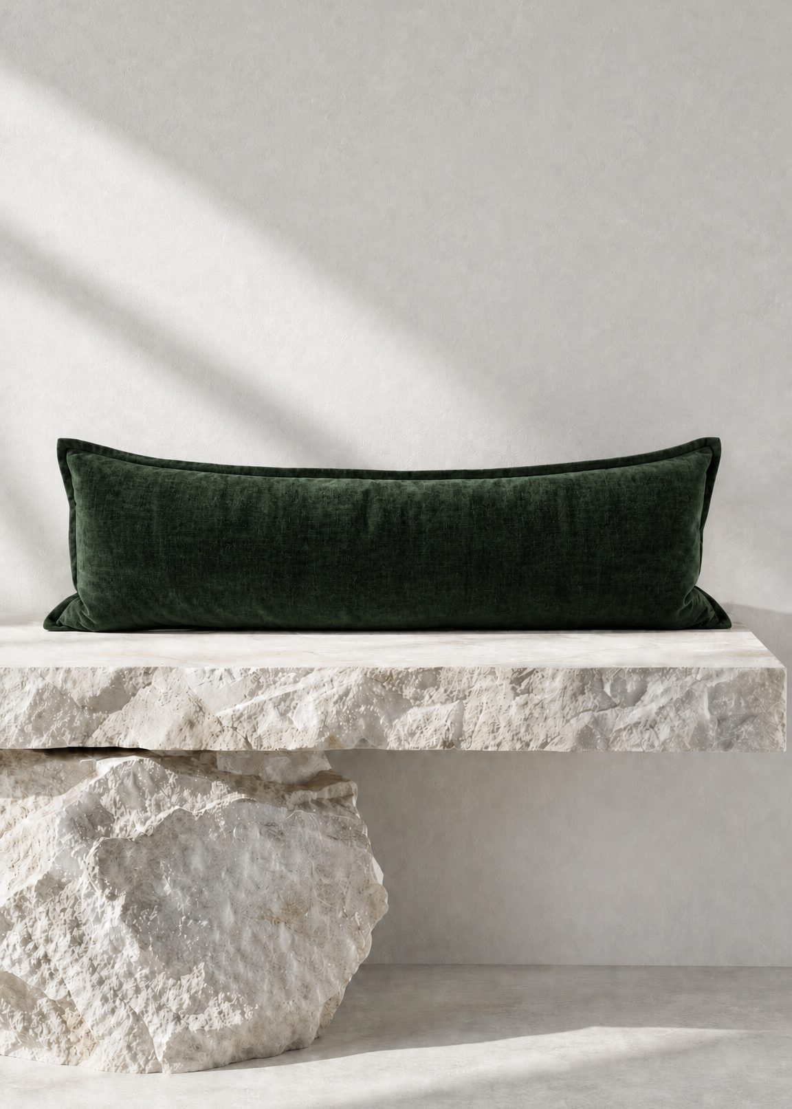 A dark forest green velvet rectangular lumbar pillow displayed on a rustic white stone bench against a textured light gray wall with soft diagonal shadows.
