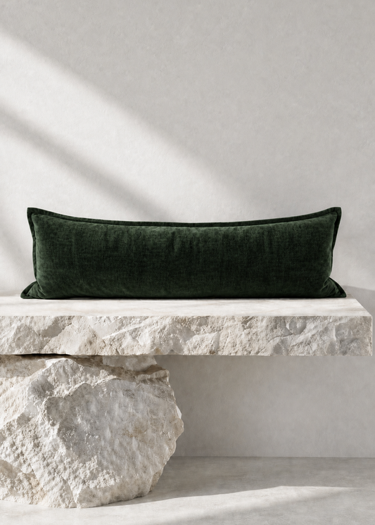 A dark forest green velvet rectangular lumbar pillow displayed on a rustic white stone bench against a textured light gray wall with soft diagonal shadows.