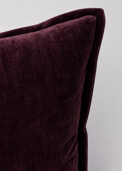 A close-up of a deep burgundy velvet throw pillow corner, showcasing its rich texture and soft piping against a neutral light gray background.