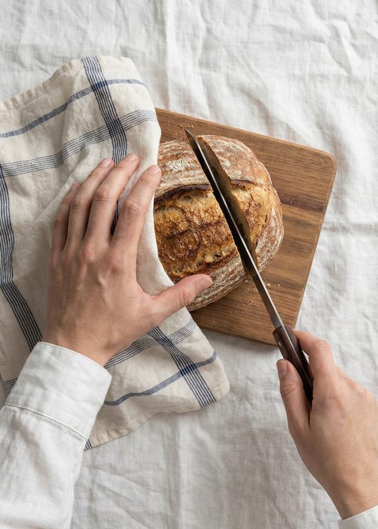 Person slicing bread on a wooden cutting board with a knife, on a white surface.