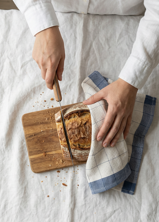Person slicing bread on a wooden cutting board with a towel nearby on a white surface