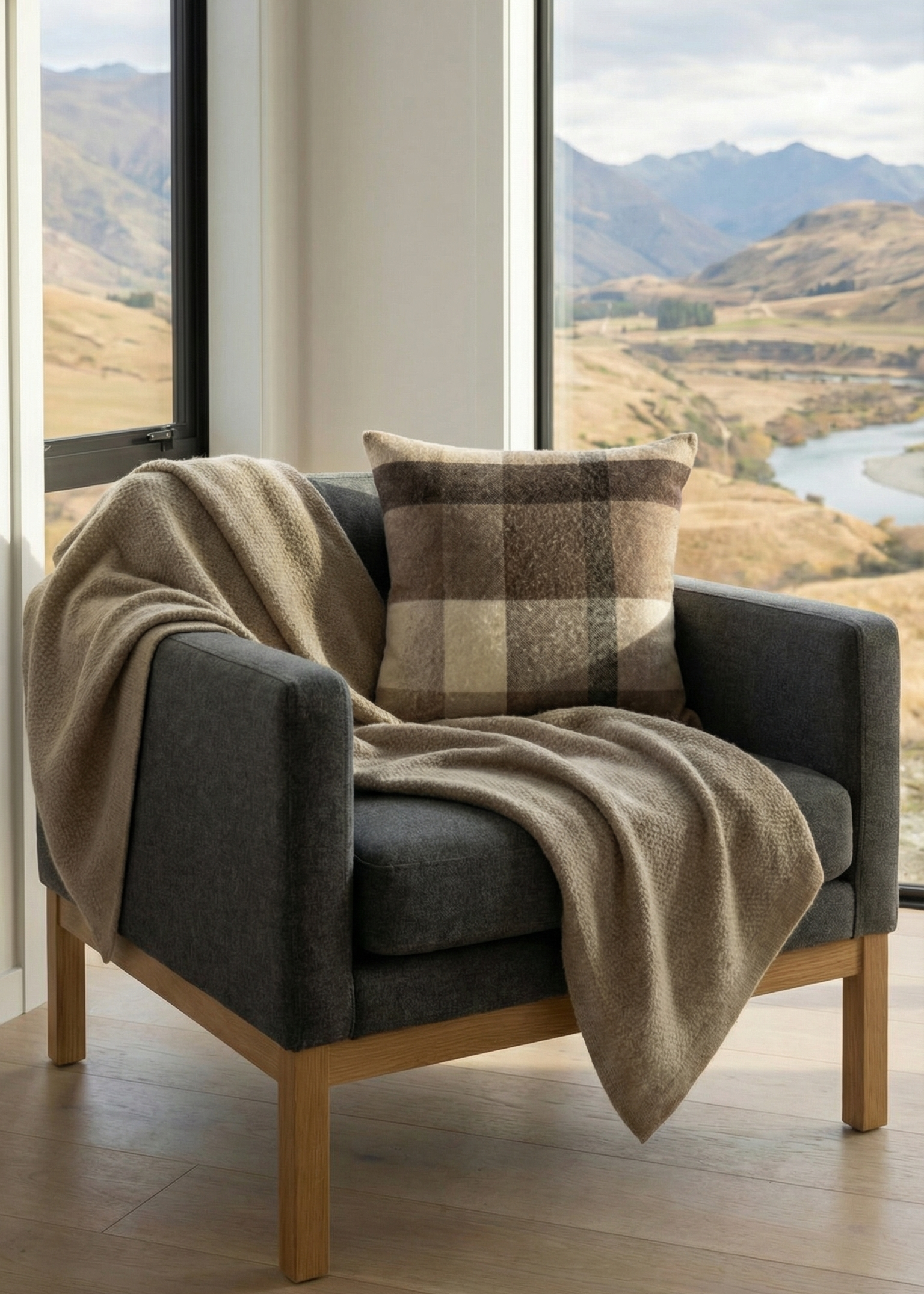 Gray armchair with a plaid cushion and blanket in a room with large windows showing a scenic view of mountains and water.