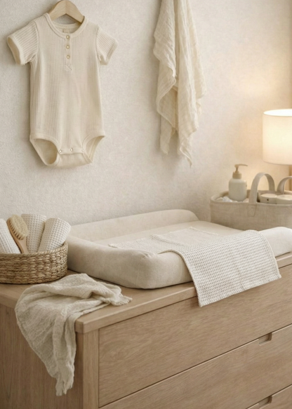 Nursery with wooden changing table, baby clothes, and lamp.