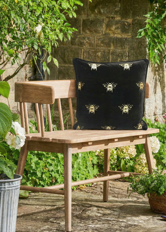 Wooden bench with a black pillow featuring bee patterns in a garden setting.