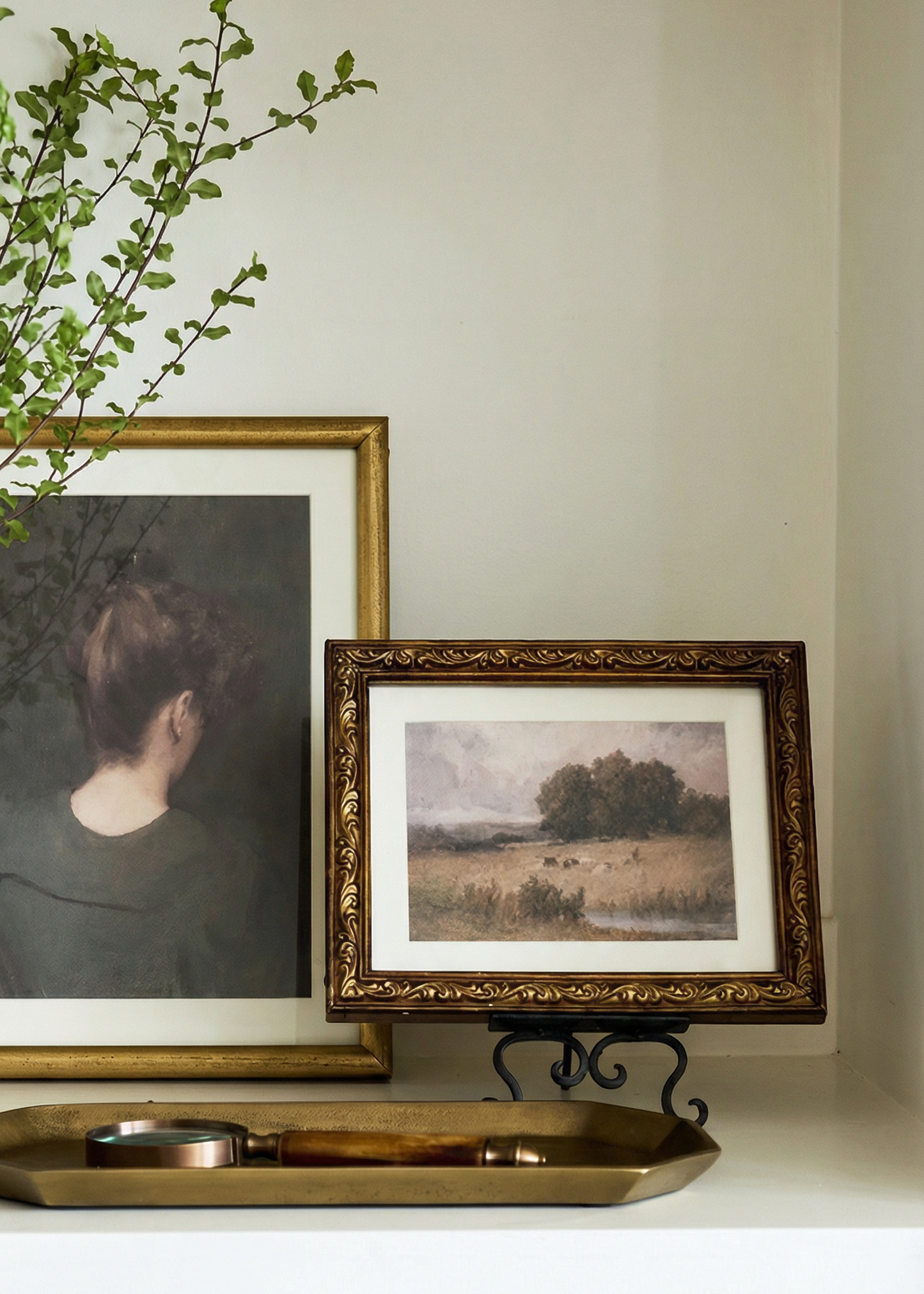 Two framed artworks on a shelf with a decorative tray and plant.