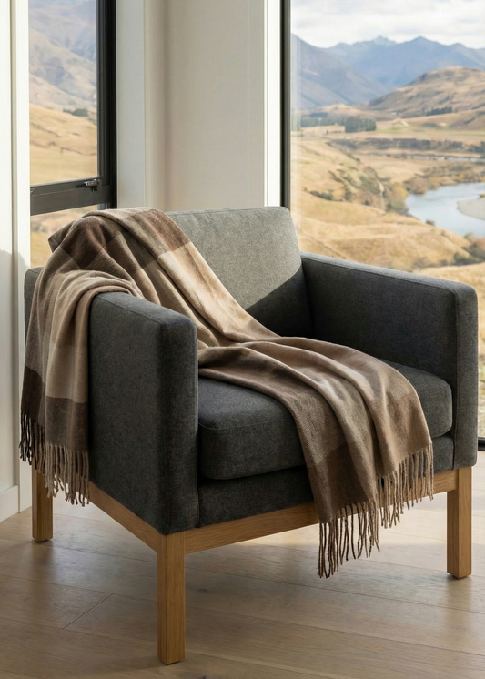 Gray armchair with a plaid blanket in a room with large windows showing a scenic view of mountains and a lake.