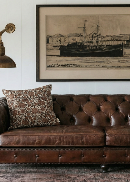 Brown leather sofa in a living room with a framed picture of a ship on the wall.