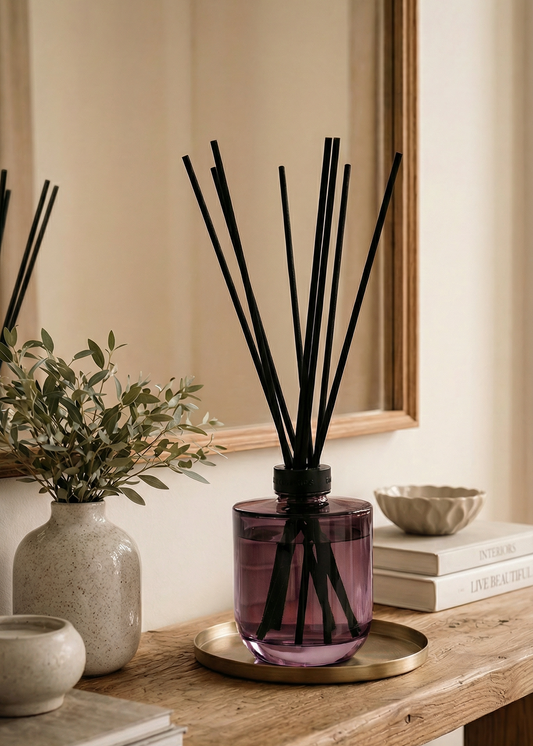 A diffuser with black sticks in a purple container on a wooden surface, surrounded by books and a vase.