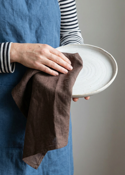 Person holding a white plate and brown towel, wearing a blue apron and striped shirt.