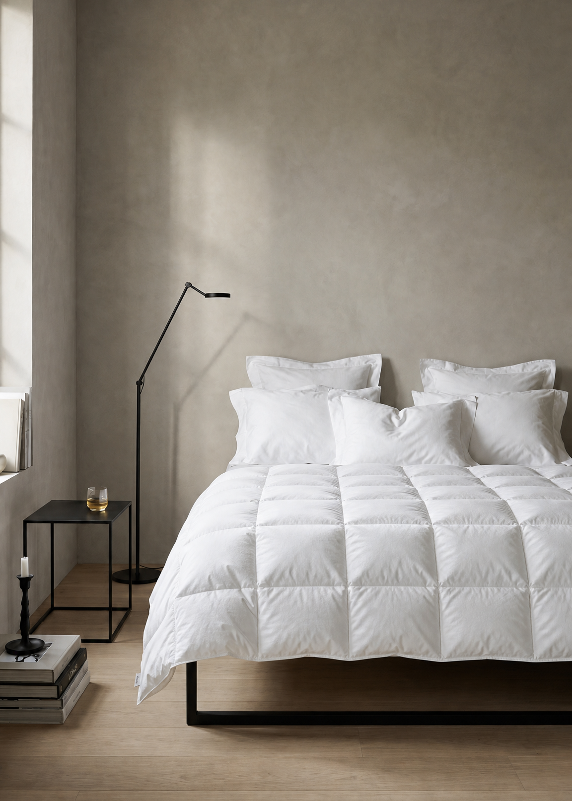 A white quilted comforter with matching white pillows on a bed in a minimalist bedroom with a textured beige wall, light wood floor, and a black floor lamp.