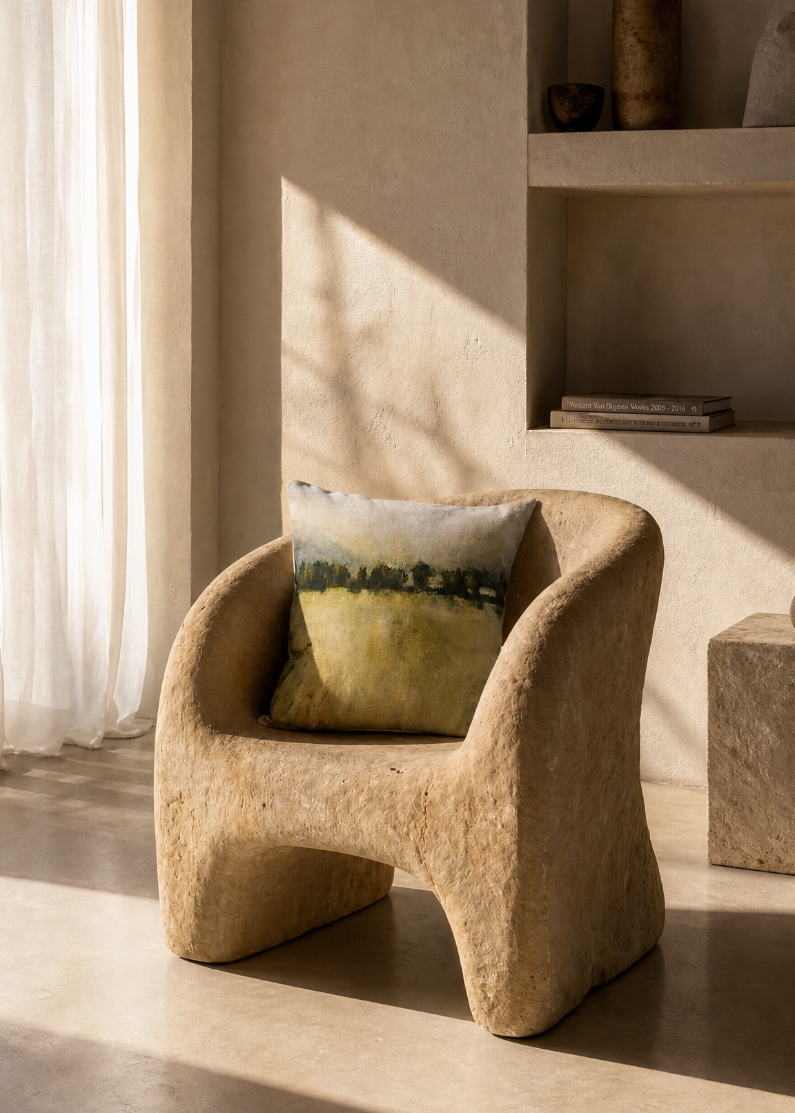 A sculptural, rounded armchair in a textured beige stone or concrete finish, styled with a green and yellow patterned accent pillow in a warm, minimalist interior setting with natural sunlight.