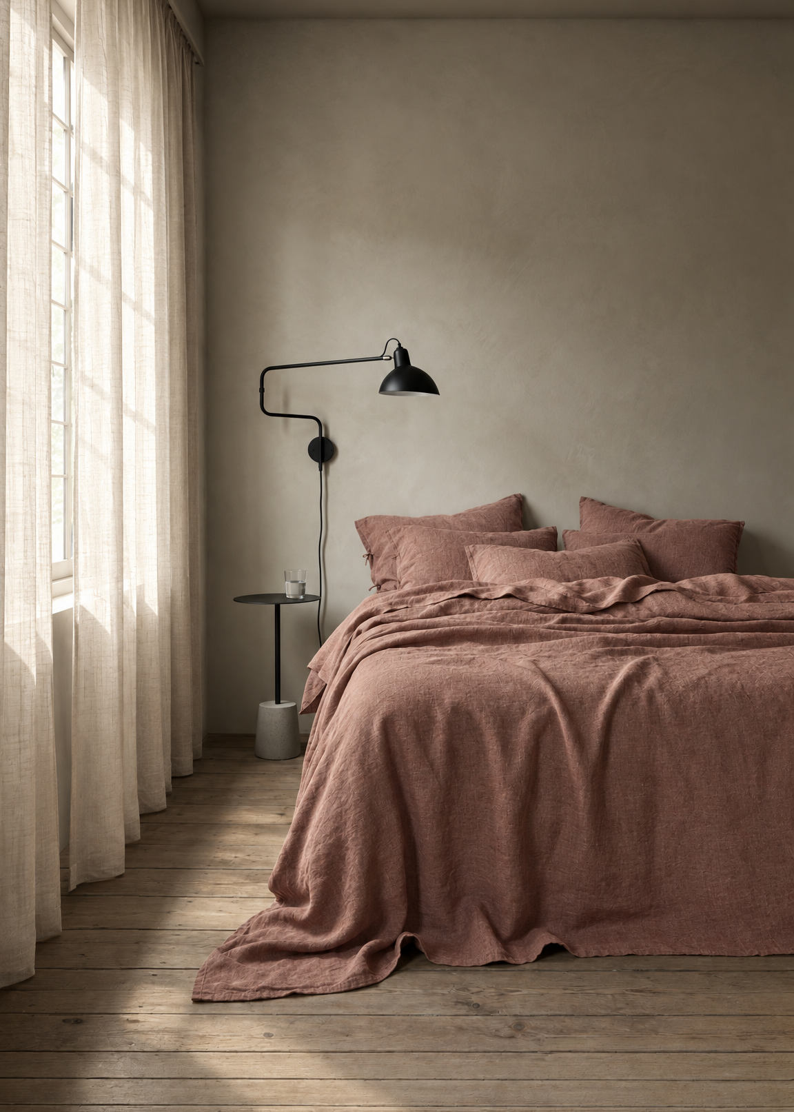 A rustic bedroom scene featuring a bed dressed in dusty rose linen bedding, set against a textured plaster wall with a black wall lamp and sheer curtains on a wooden floor.