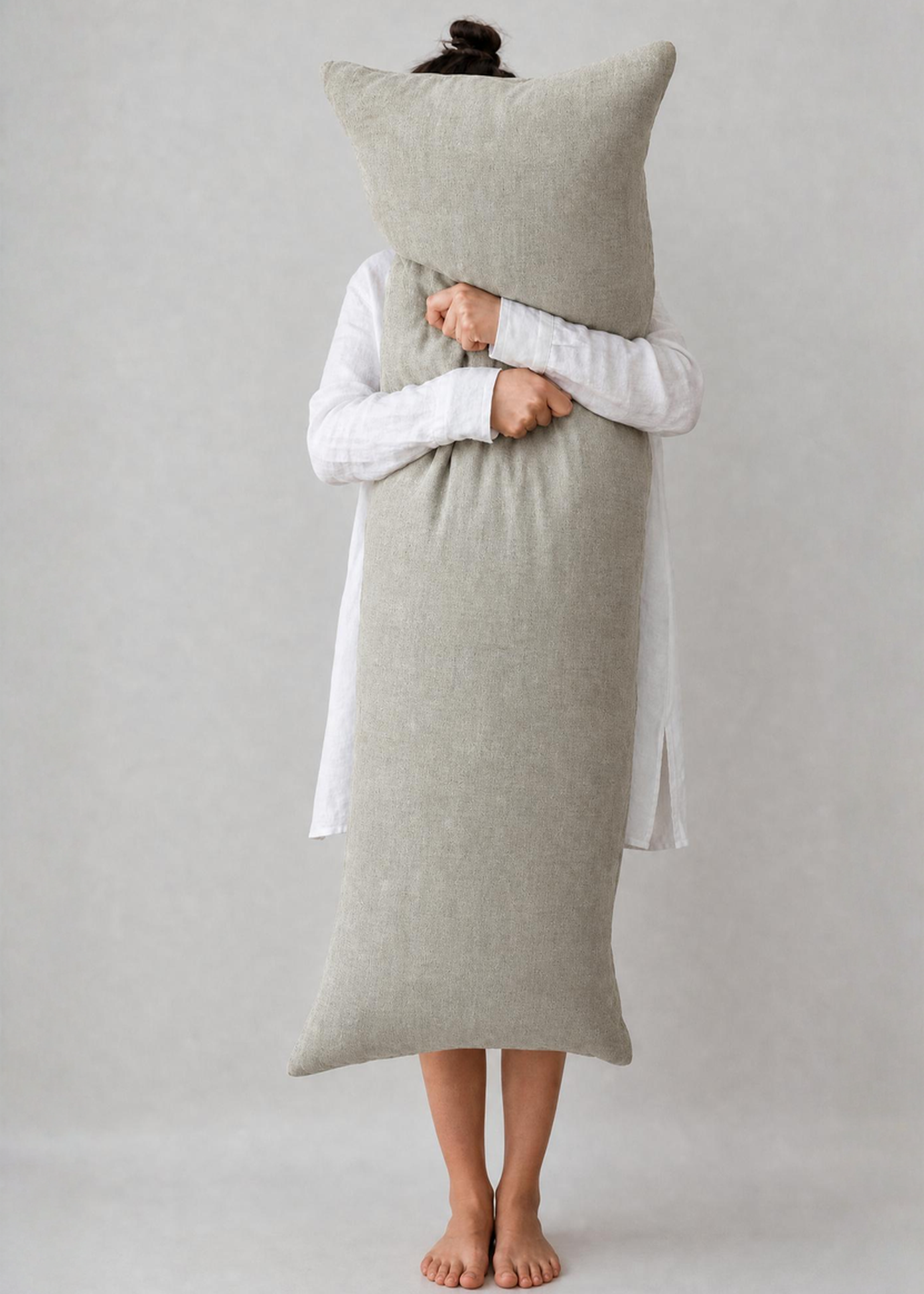 A person standing barefoot against a light gray studio background, wrapped in a long, oversized light beige pillow or blanket and holding a matching square pillow in front of their face. They are wearing a white long-sleeve top.