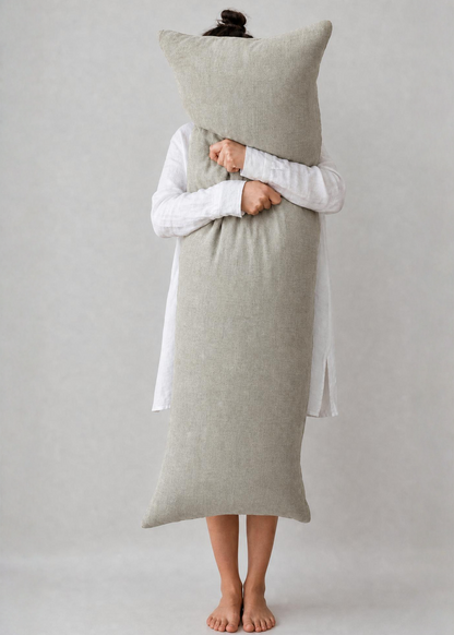 A person standing barefoot against a light gray studio background, wrapped in a long, oversized light beige pillow or blanket and holding a matching square pillow in front of their face. They are wearing a white long-sleeve top.
