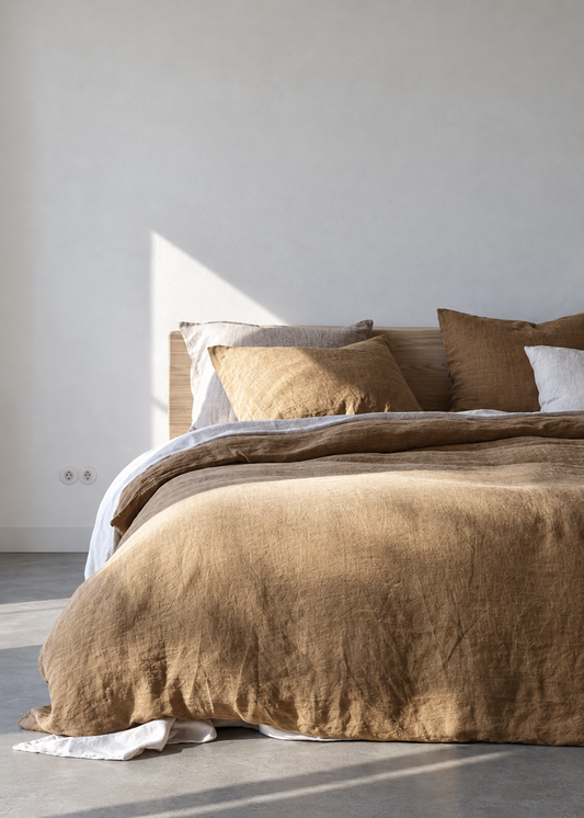 A bed dressed in earthy brown and beige linens with matching pillows, styled in a minimalist bedroom with a concrete floor and a sunlit white wall.