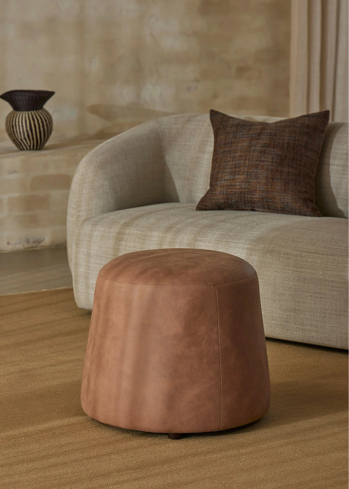 A round, tapered brown leather ottoman in the foreground, set on a textured beige rug in front of a curved light beige sofa with a dark brown accent pillow.