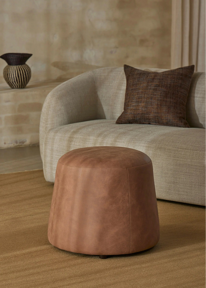 A round, tapered brown leather ottoman in the foreground, set on a textured beige rug in front of a curved light beige sofa with a dark brown accent pillow.