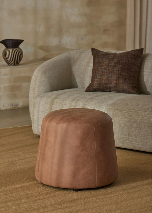 A round, tapered brown leather ottoman in the foreground, set on a textured beige rug in front of a curved light beige sofa with a dark brown accent pillow.