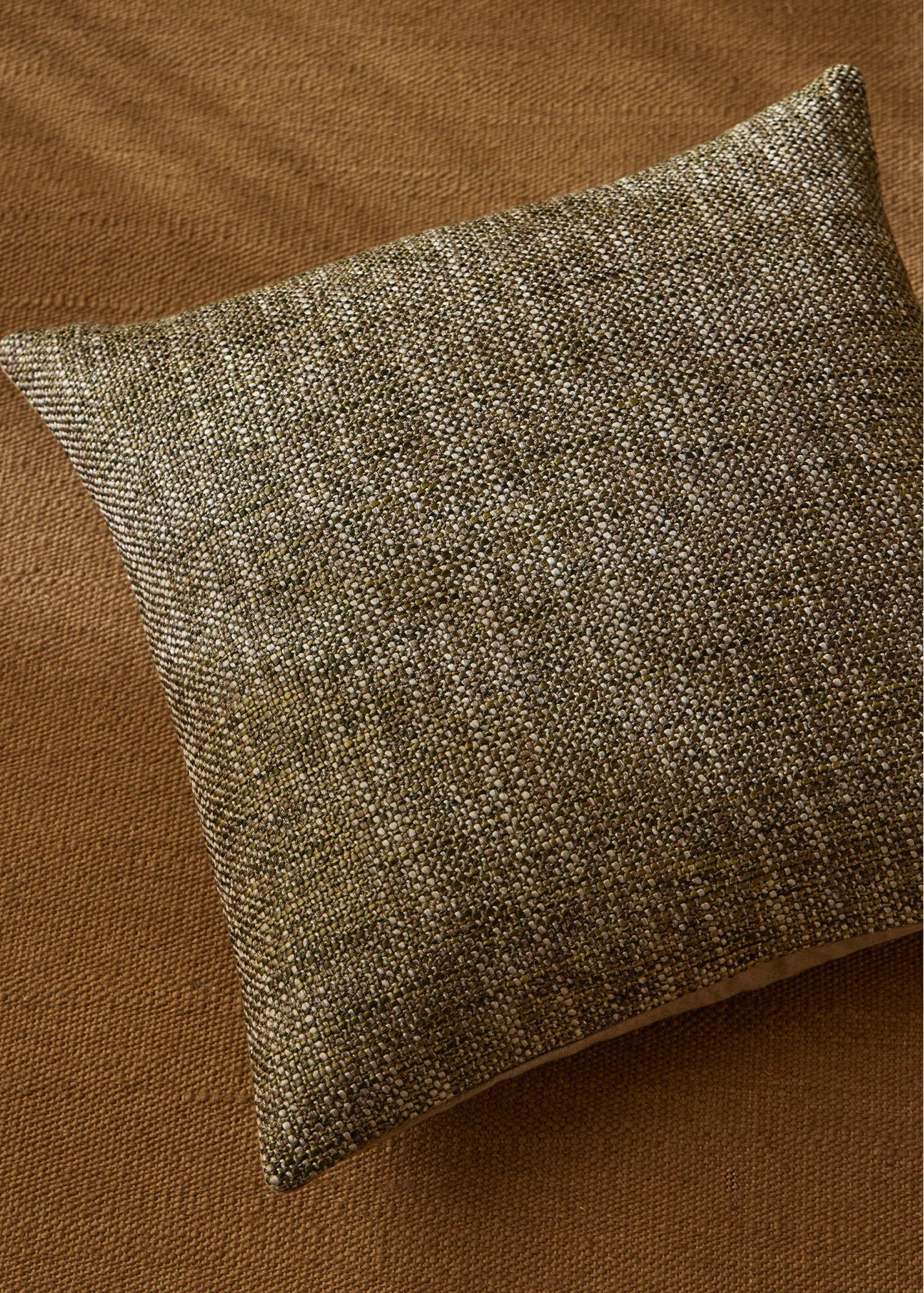 A square throw pillow with a textured woven fabric in a blend of olive green, brown, and beige, resting on a matching brown textured surface.