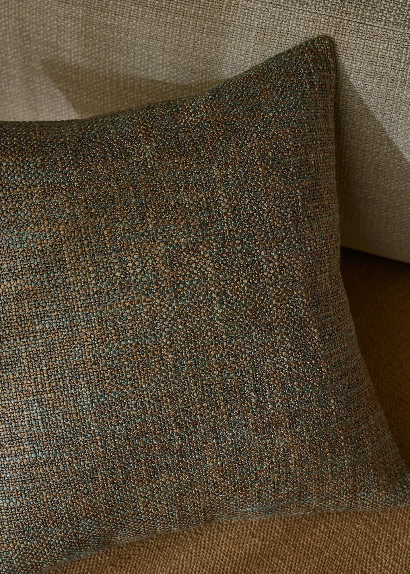 A square throw pillow with a textured, woven herringbone pattern in earthy brown, beige, and muted blue-gray tones, resting on a matching woven sofa.