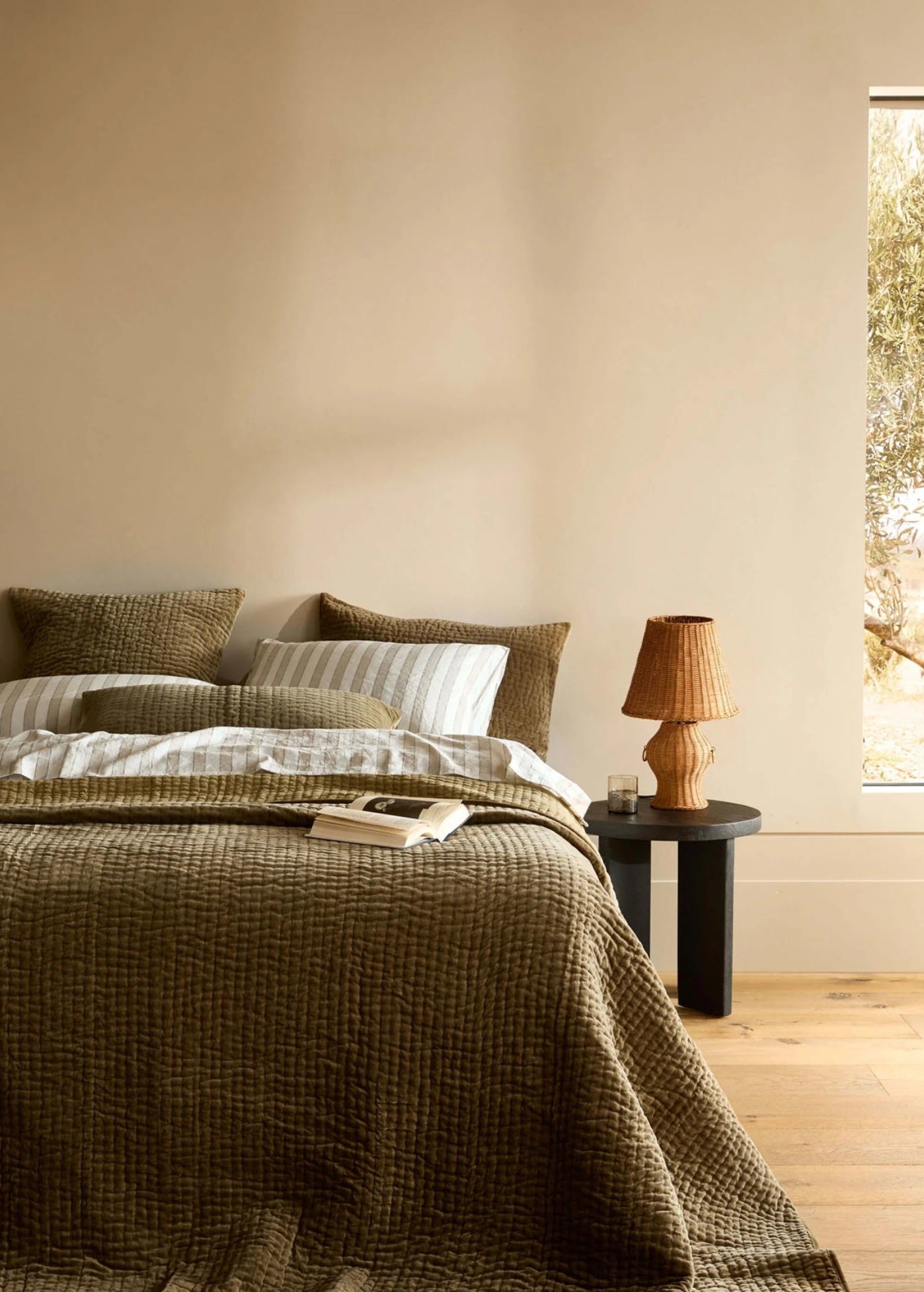Bedroom with a bed covered in a green blanket, a lamp on a nightstand, and a window showing trees.
