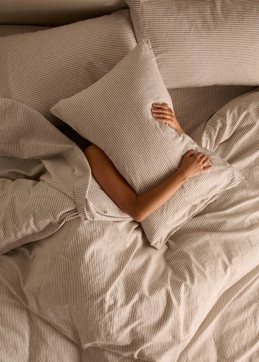 Person lying in bed with beige bedding and a pillow, partially covered by the blanket.