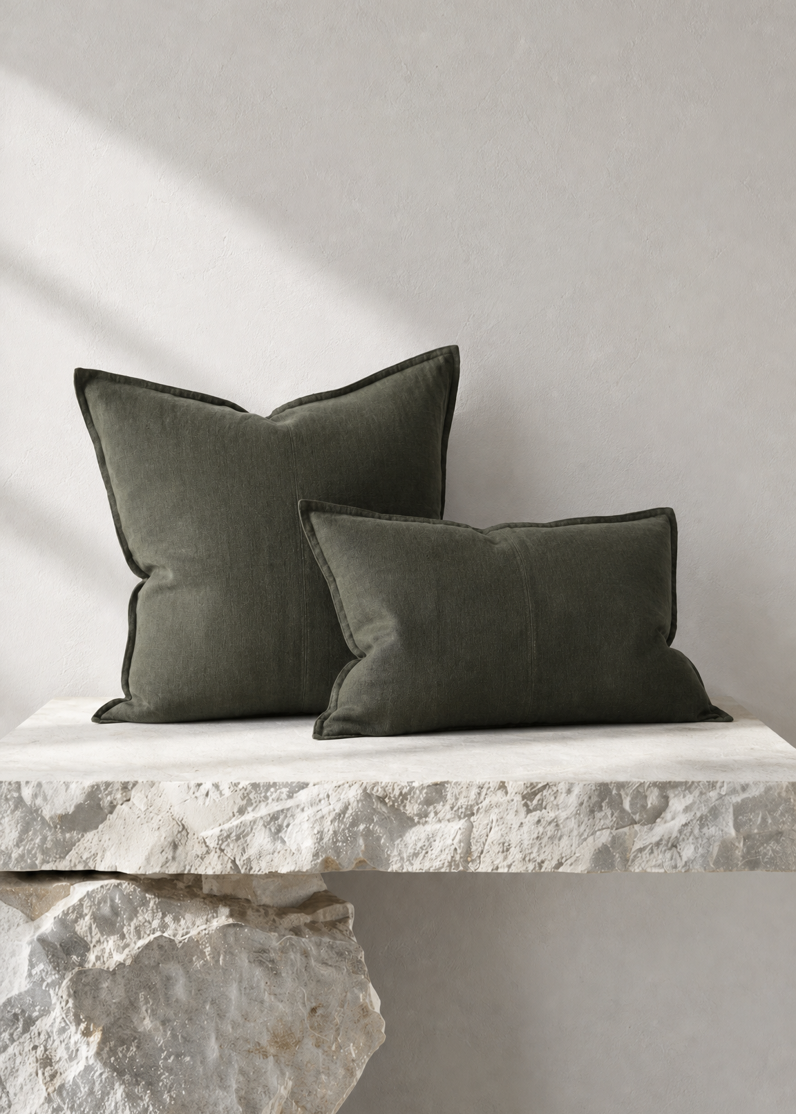 Two dark olive green throw pillows displayed on a rustic white stone bench against a textured light gray wall.