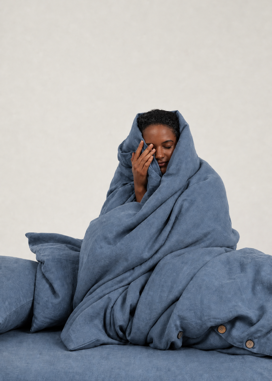 A person wrapped in a dusty blue blanket sits on a matching blue bed against a light beige wall.