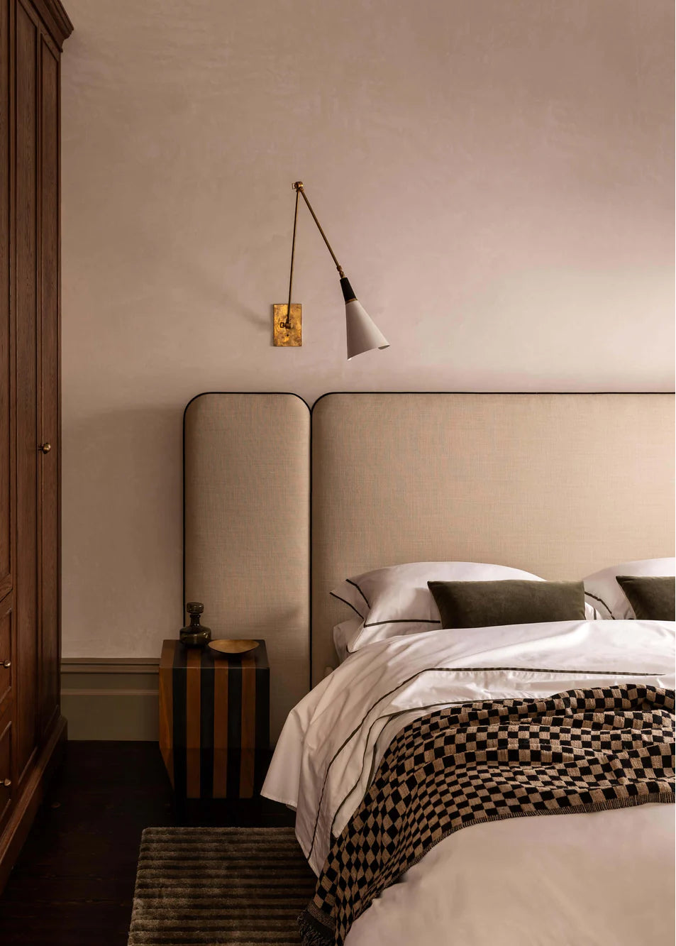 The image depicts a sophisticated bedroom setting featuring a beige olive-colored duvet cover with black grosgrain ribbon trim detailing. A white lamp with a gold mount is mounted on a wall on the left, providing a contrast in color and texture.
