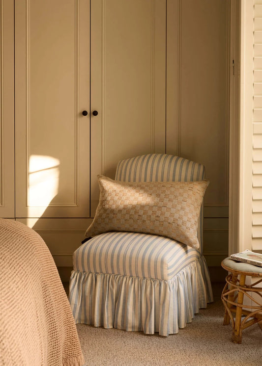 A cozy bedroom corner featuring a ruffled slipcovered armchair with vertical stripes in beige and cream, styled with a matching patterned throw pillow. A large beige paneled wardrobe stands in the background, with warm sunlight casting soft shadows on the wall and floor.