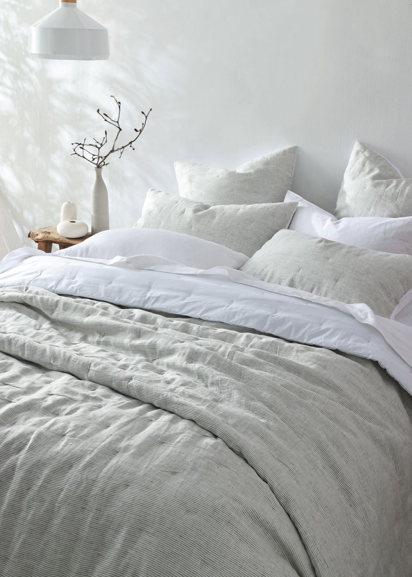 A neatly made bed with light gray and white bedding, including a textured gray duvet cover and matching pillows, styled in a bright, minimalist bedroom with a white wall, hanging lamp, and small side table.