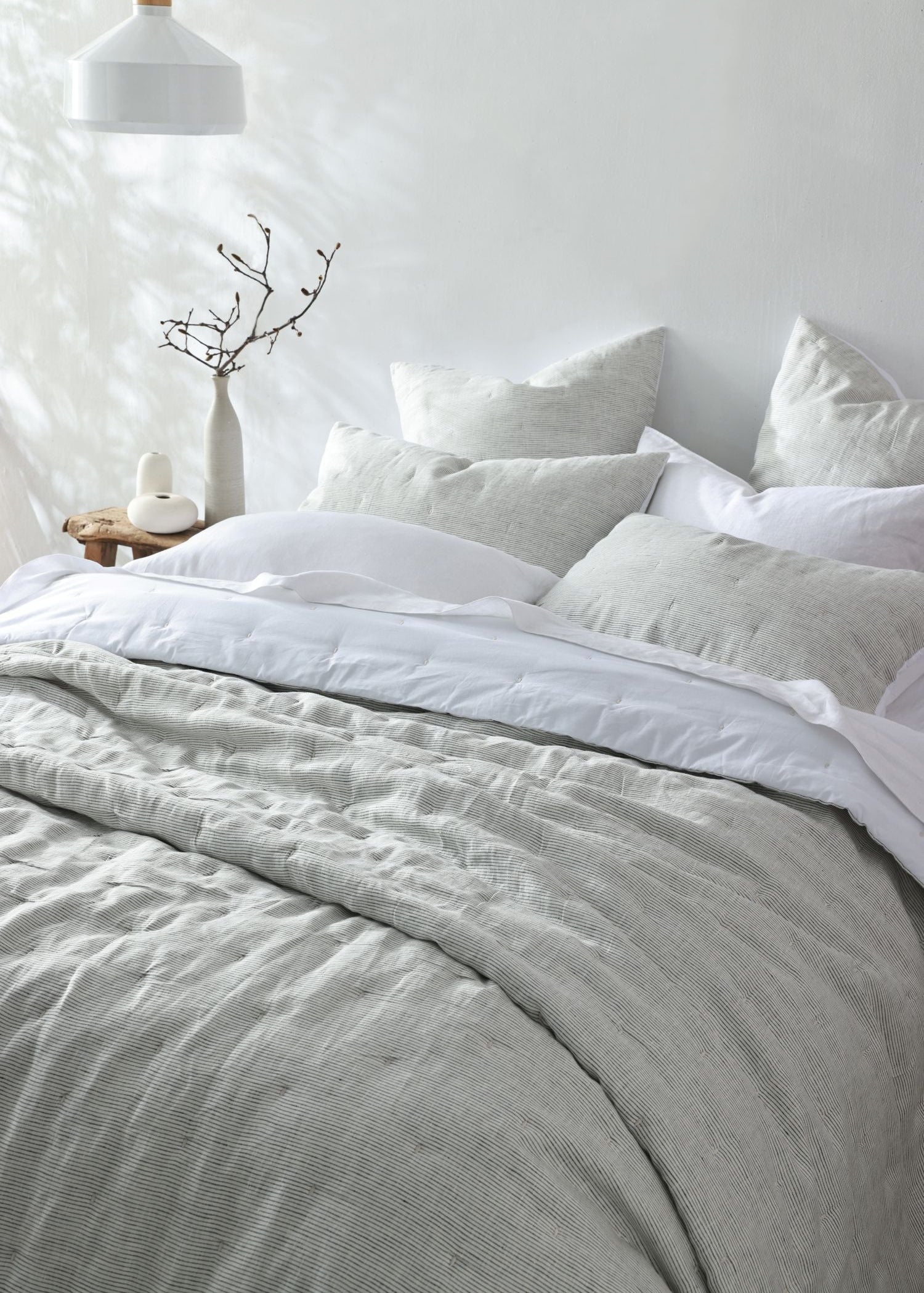 A neatly made bed with light gray and white bedding, including a textured gray duvet cover and matching pillows, styled in a bright, minimalist bedroom with a white wall, hanging lamp, and small side table.
