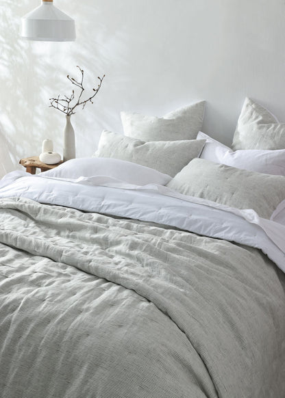 A neatly made bed with light gray and white bedding, including a textured gray duvet cover and matching pillows, styled in a bright, minimalist bedroom with a white wall, hanging lamp, and small side table.