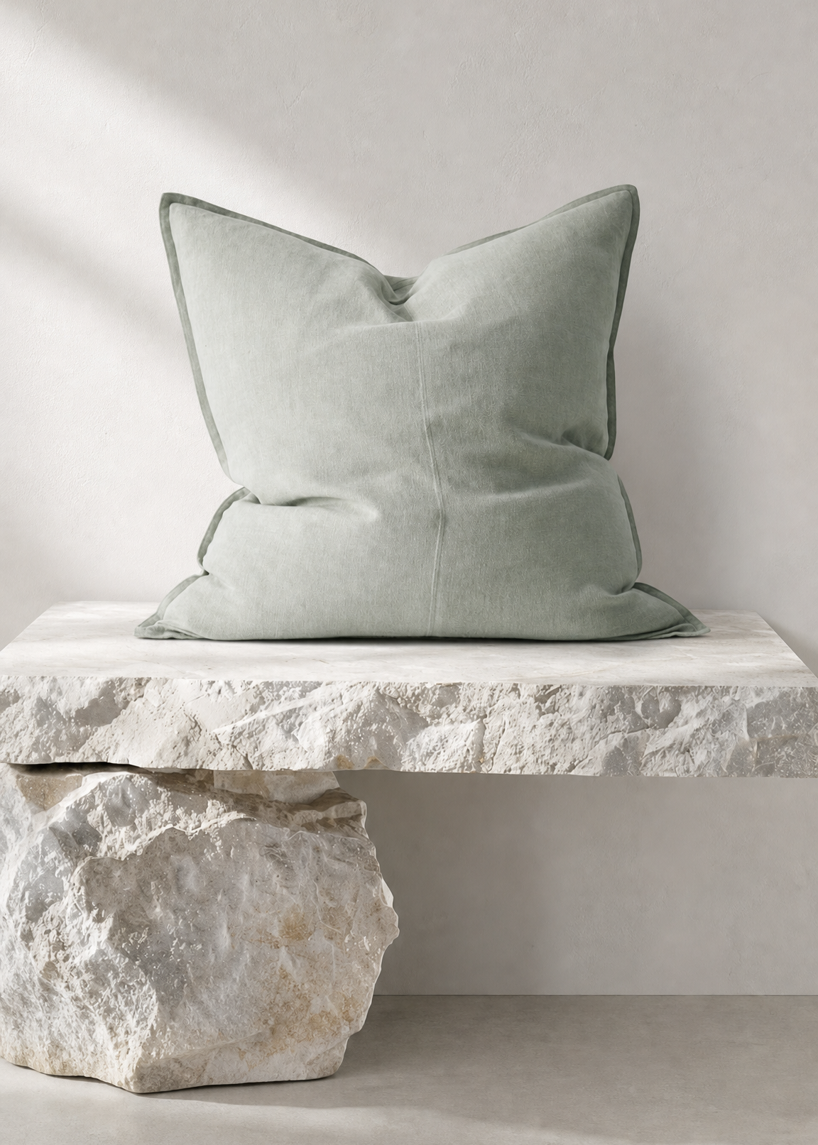 A square sage green throw pillow with piped edges, resting on a white stone bench against a neutral light gray wall.