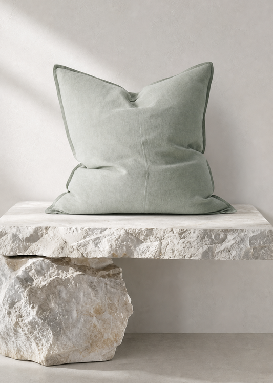 A square sage green throw pillow with piped edges, resting on a white stone bench against a neutral light gray wall.