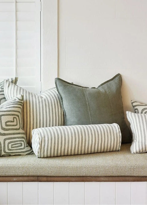 A built-in bench seat with a textured beige cushion and multiple decorative throw pillows, including striped and geometric patterned pillows, set against a white paneled wall and shutters.