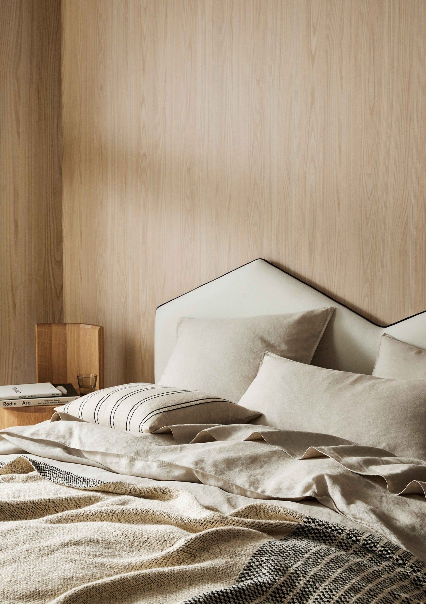 A bedroom scene with a neutral-colored linen pillowcase on the bed, alongside a wooden headboard and bedside table with a lamp.